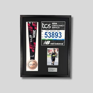 May include: A black framed shadow box display containing a TCS New York City Marathon medal, race bib with number 53893, and a small photo of a runner. The medal has a red and black ribbon. The frame is on a light grey background.