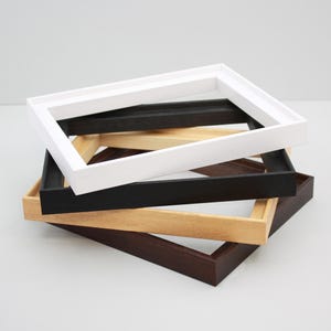May include: A stack of four rectangular picture frames in varying finishes. The top frame is white, followed by a black frame, a light natural wood frame, and a dark brown wood frame at the bottom. Each frame features a simple, clean profile.