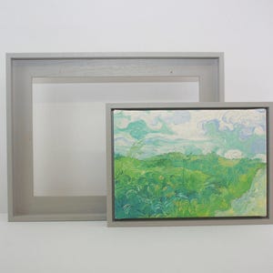 May include: A framed landscape painting with a larger, empty frame behind it. The painting shows a green and blue landscape with a cloudy sky. The painting is rectangular and has a canvas texture. The frames are grey.