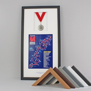 May include: Framed artwork featuring a Manchester Marathon map, a medal, and a red ribbon. The map is blue with a red route and white text. A selection of wooden frame samples in various colours are displayed in front of the framed artwork.