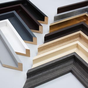May include: A selection of wooden picture frames in shades of white, grey, black, and brown. The frames are displayed at angles, highlighting their different profiles and finishes. Some frames show visible wood grain.