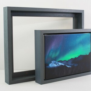 May include: Two framed canvas art pieces. One is a large, empty, dark grey frame. The other is a smaller canvas with a vibrant image of the Northern Lights, featuring green, blue, and purple hues over a mountain range.