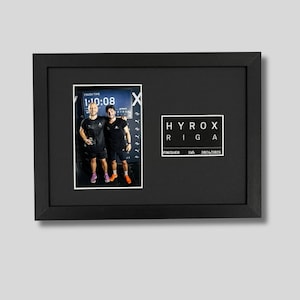 May include: A black framed photo collage with two images. The left image shows two people in athletic wear, with a timer displaying 1:10:08. The right image has the text "HYROX RIGA" and "FINISHER LVA 2024/2025" in a white box. The frame is black.