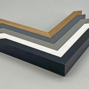 May include: A selection of wooden picture frame samples in a range of colours: black, white, grey, and natural wood. The frames are arranged in a layered, angled display, showing their different profiles and finishes. These frames are designed for artwork or photographs.