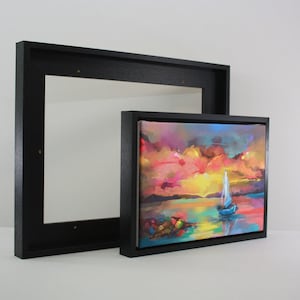 May include: A framed canvas artwork depicting a sunset with a sailboat, in a black frame. Beside it is a larger, empty black frame. The painting uses vibrant colours including pink, orange, yellow, and blue. The artwork is approximately 20 cm x 30 cm.