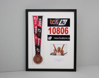 May include: Framed black picture frame with a white mat. The frame holds a running medal with a red and black ribbon, a race bib with the number 10806, a New Balance logo, and a small photo of runners.
