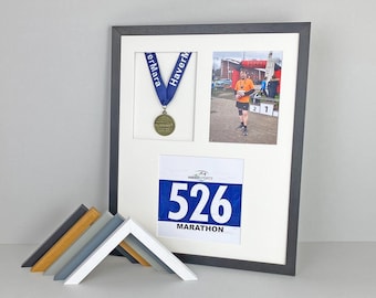 May include: A black rectangular display frame with a white mat containing a blue medal ribbon, a circular medal, a photograph of a runner, and a race bib with the number 526 and the text MARATHON. Several small frame corner samples are in front.