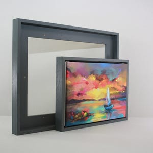 May include: A small, square painting of a sailboat on the water, framed in a dark grey wooden frame. The painting features vibrant colours of pink, yellow, and blue. A larger, empty dark grey frame is behind the painting.