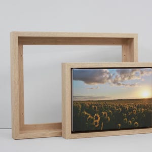 May include: Two wooden picture frames, one empty and one with a canvas print. The print shows a field of sunflowers at sunset, with a bright yellow sun and a cloudy sky. The frames are made of light-coloured wood.