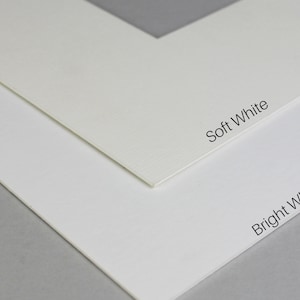 Puede incluir: Primer plano de dos cartulinas blancas, una etiquetada como "Soft White" y la otra como "Bright White". Las cartulinas son texturizadas y muestran una sutil diferencia de color.