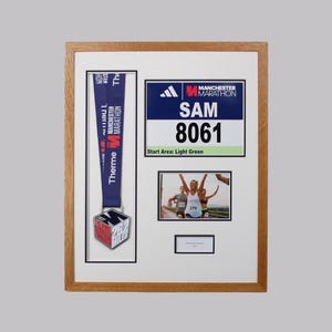 May include: A wooden frame containing a Manchester Marathon medal with a blue ribbon, a race bib for Sam number 8061, a photograph of runners, and a small blank nameplate. The display features white matting and a clear layout for memorabilia.