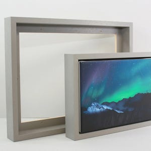 May include: A framed canvas print displaying the aurora borealis. The artwork shows a mountain range beneath a night sky with green, blue, and purple tones. The frame is a light grey colour.