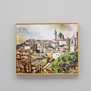 May include: A framed canvas print depicting a cityscape with a church in the background. The buildings are painted in warm tones of brown, yellow, and orange. The sky is a soft blue with white clouds. The print is framed in a light wood frame.