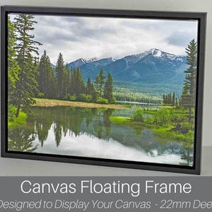 May include: A framed canvas print depicting a landscape with a lake, trees, and mountains under a cloudy sky. The canvas is set in a black floating frame, 22mm deep. The image is a nature scene with reflections in the water.