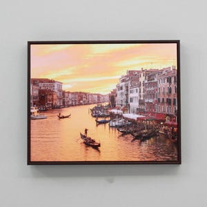 May include: A framed photograph depicting a canal in Venice, Italy, with gondolas and buildings lining the waterway. The sky is a vibrant orange and pink, creating a warm and inviting atmosphere.