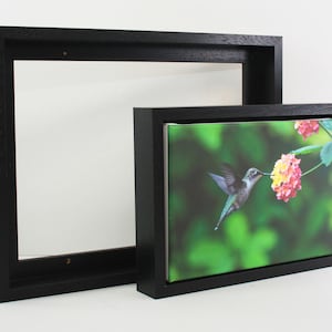 May include: Two black wooden picture frames, one empty and one with a canvas print. The print shows a hummingbird with iridescent feathers feeding on a flower with pink and yellow blossoms against a green backdrop.