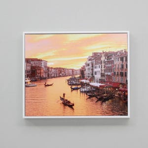 May include: A framed photograph of a canal in Venice, Italy, at sunset. The canal is lined with buildings and there are gondolas in the water. The sky is a vibrant orange and pink.