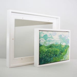 May include: Two white wooden picture frames, one empty and the other holding a landscape painting. The painting depicts green foliage and a blue sky with clouds. The frames are rectangular.