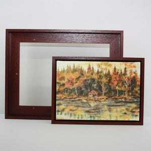 May include: Two framed landscape paintings. One is a rectangular painting of a lakeside scene with trees in autumn colours. The other is a larger, empty, rectangular frame. Both frames are dark brown.