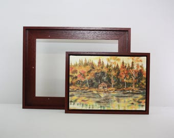 Puede incluir: Dos pinturas de paisajes enmarcadas. Una es una pintura rectangular de una escena junto al lago con árboles en colores otoñales. La otra es un marco rectangular más grande y vacío. Ambos marcos son de color marrón oscuro.