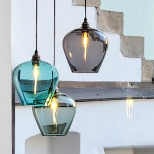 May include: Three pendant lights with glass shades in teal and grey hang from a white ceiling fixture. The lights are suspended by black cords, illuminating a white wall and stone staircase. The glass shades have a teardrop shape.