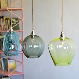 May include: Three glass pendant lights in shades of blue, gray, and green hang from brown cords. The lights have a bulb inside and are suspended against a neutral wall. A pink bookshelf filled with books is visible in the background.