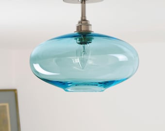 May include: A modern ceiling light fixture with a brushed nickel finish and a large, oval-shaped, turquoise glass shade. The light bulb is visible inside the shade. A framed picture is in the background.
