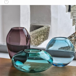 May include: Three glass vases in aubergine, emerald, and turquoise hues. The aubergine vase is tall and rounded, the emerald vase is low and wide, and the turquoise vase is spherical. The vases are displayed on a wooden surface with the text "OPTION 6".