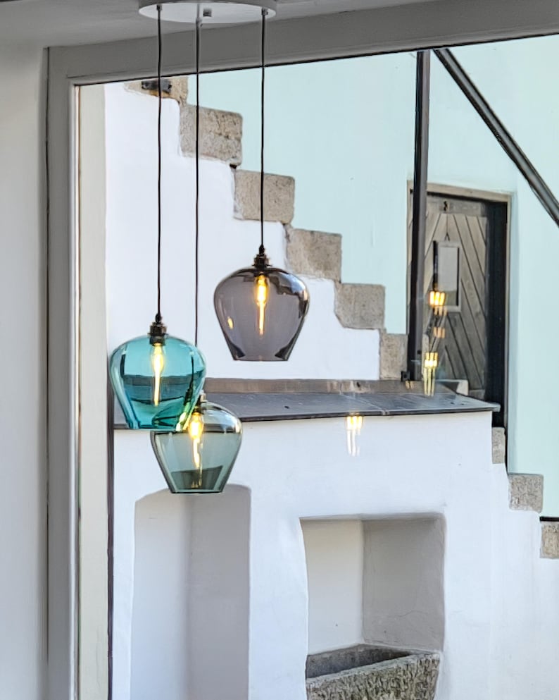 May include: Three pendant lights with glass shades in teal and grey hang from a white ceiling fixture. The lights are suspended by black cords, illuminating a white wall and stone staircase. The glass shades have a teardrop shape.