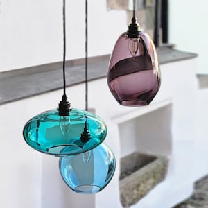 May include: Three hanging glass pendant lights in shades of teal, blue, and purple. Each light has a unique shape, with a bulb visible inside. The lights are suspended by black cords against a white background.