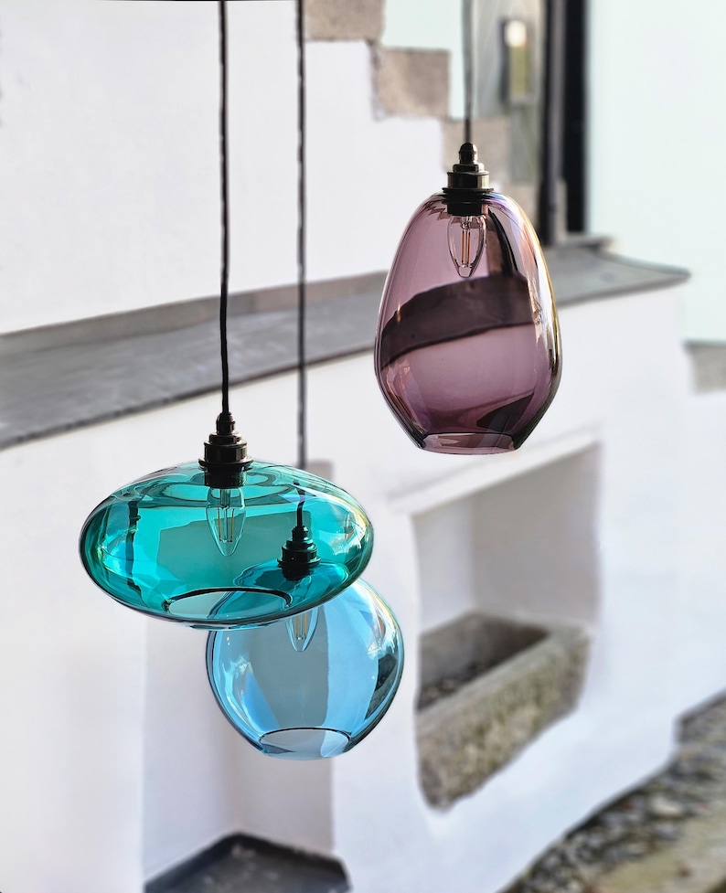 May include: Three hanging glass pendant lights in shades of teal, blue, and purple. Each light has a unique shape, with a bulb visible inside. The lights are suspended by black cords against a white background.