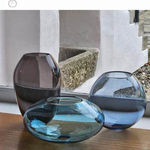 May include: Three glass vases in shades of gray, turquoise, and blue are displayed. The vases have rounded shapes, with the turquoise vase being the flattest. The text "OPTION 5" is visible. The vases are on a wooden surface.
