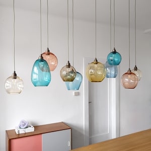 Puede incluir: Un conjunto de ocho lámparas colgantes de vidrio que cuelgan del techo. Las lámparas son de varios tonos de azul, amarillo y rosa.