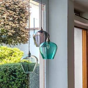 May include: Three pendant lights with glass shades in clear, grey, and teal green hang in front of a window. The shades have a unique, rounded shape and are suspended by black cords. The lights are turned on.