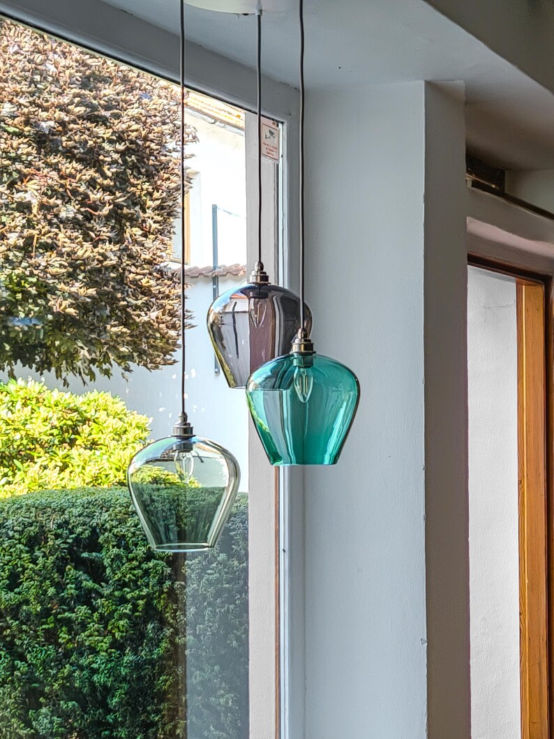 May include: Three pendant lights with glass shades in clear, grey, and teal green hang in front of a window. The shades have a unique, rounded shape and are suspended by black cords. The lights are turned on.