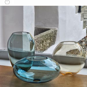 May include: Three glass vases in smoke grey, turquoise, and whiskey colors. The vases have unique shapes: a tall, rounded vase, a flattened oval, and a spherical vase. The text "OPTION 10" is at the top, and the text "SMOKE GREY", "TURQUOISE", and "WHISKEY" are at the bottom.