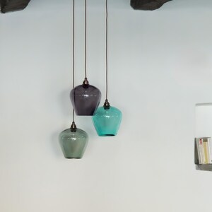 May include: Three glass pendant lights in shades of grey, teal, and green hang from the ceiling. Each light has a rounded, vase-like shape with a dark cord and a metal fixture. The lights are suspended against a white wall.