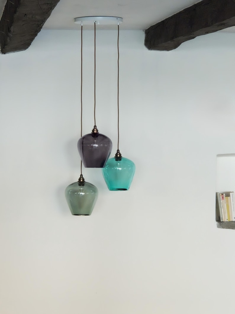 May include: Three glass pendant lights in shades of grey, teal, and green hang from the ceiling. Each light has a rounded, vase-like shape with a dark cord and a metal fixture. The lights are suspended against a white wall.