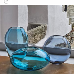 May include: Three glass vases in turquoise, aquamarine, and smoke blue hues. The vases have rounded shapes, with one being a flattened oval. The text "OPTION 12" is visible. The vases are displayed on a wooden surface.