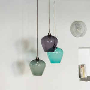 May include: Three glass pendant lights in shades of green, purple, and teal. The lights hang from brown cords attached to a dark wooden beam. The shades have a rounded, bell-like shape, and the fixtures have a metallic finish.