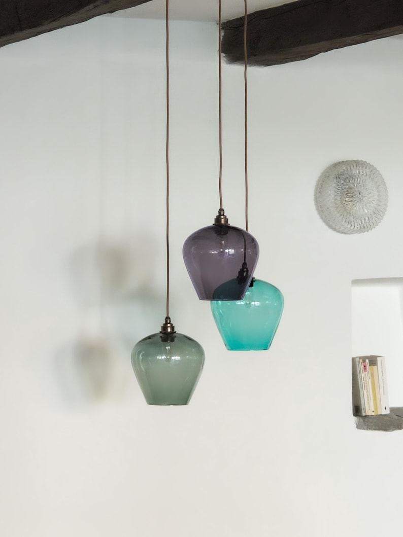 May include: Three glass pendant lights in shades of green, purple, and teal. The lights hang from brown cords attached to a dark wooden beam. The shades have a rounded, bell-like shape, and the fixtures have a metallic finish.