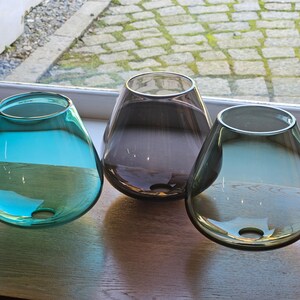 May include: Three teardrop-shaped glass vases in teal, grey, and smoky purple sit on a wooden surface. Each vase has a wide opening and a small hole near the base. The vases are transparent, allowing light to pass through.