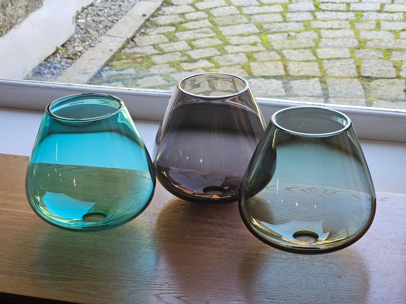 May include: Three teardrop-shaped glass vases in teal, grey, and smoky purple sit on a wooden surface. Each vase has a wide opening and a small hole near the base. The vases are transparent, allowing light to pass through.