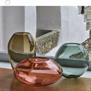 May include: Three glass vases in shades of old gold, aurora, and smoke grey sit on a wooden surface. The vases have unique shapes, including a tall, rounded vase, an oval-shaped vase, and a spherical vase. The text "OPTION 8" is visible.