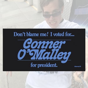 May include: A black and white sign with the text "Don't blame me! I voted for... Conner O'Malley for president. slopcorp.lol" in white text. The text "Conner O'Malley" is in a blue, stylized font.
