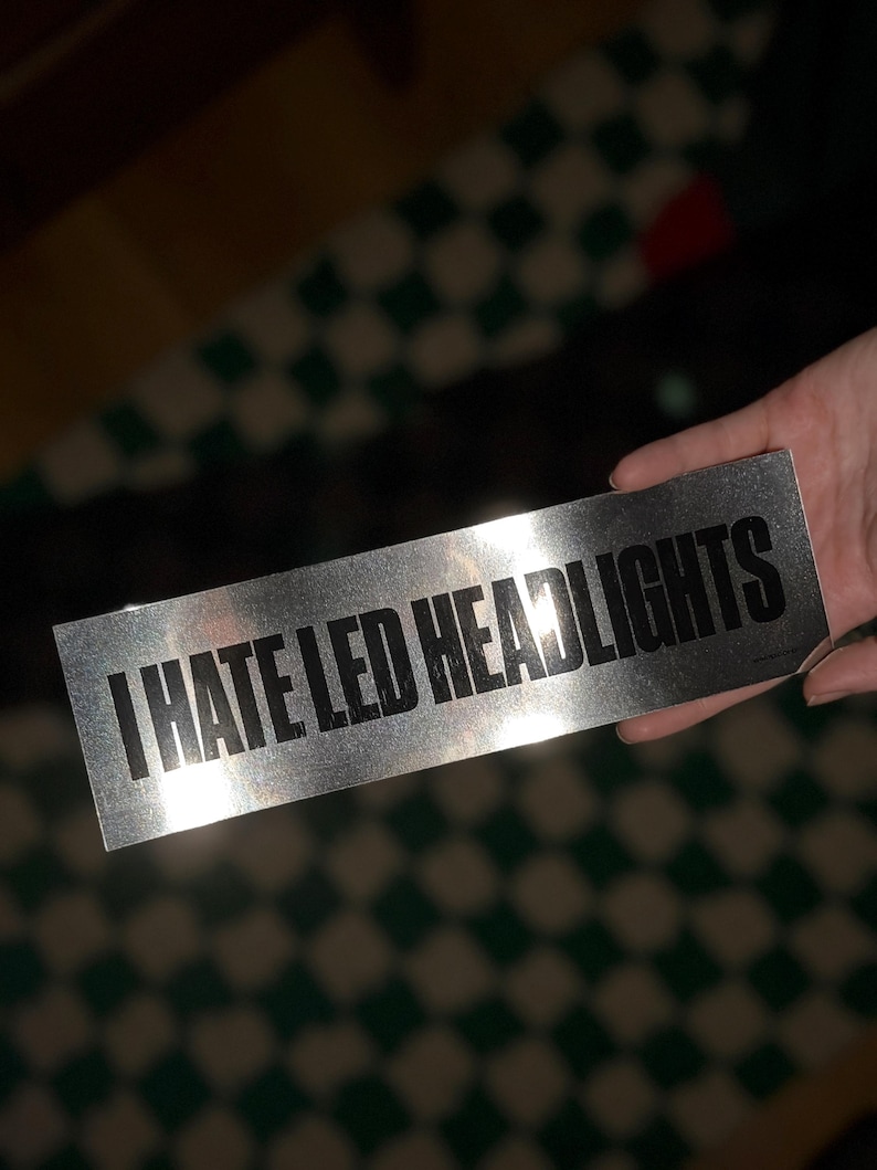 以下が含まれることがあります： 「I HATE LED HEADLIGHTS」のテキストが黒い大文字で書かれた長方形の銀色のステッカー。ステッカーは、緑と白のチェック柄のぼやけた背景に対して手で持たれています。