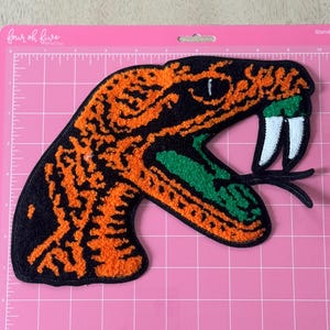 May include: Embroidered patch featuring a snake head with an open mouth. The snake has orange and black markings, green inner mouth, white fangs, and a black tongue. The patch is on a pink cutting mat.