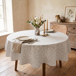 May include: A round dining table set for a meal. The table is covered with a light grey tablecloth with a white floral pattern. A vase of flowers, candles, plates, and silverware are arranged on the table.