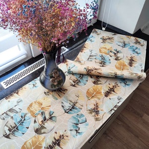 May include: A beige table runner with a leaf pattern in shades of blue, brown, and yellow. The runner is folded in half and is sitting on a dark countertop. A vase with dried flowers is in the background.