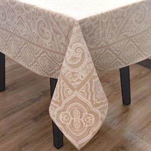 May include: A beige tablecloth featuring a detailed paisley pattern. The fabric is draped over a table, highlighting the intricate design. The colour scheme is neutral, with shades of beige and cream.